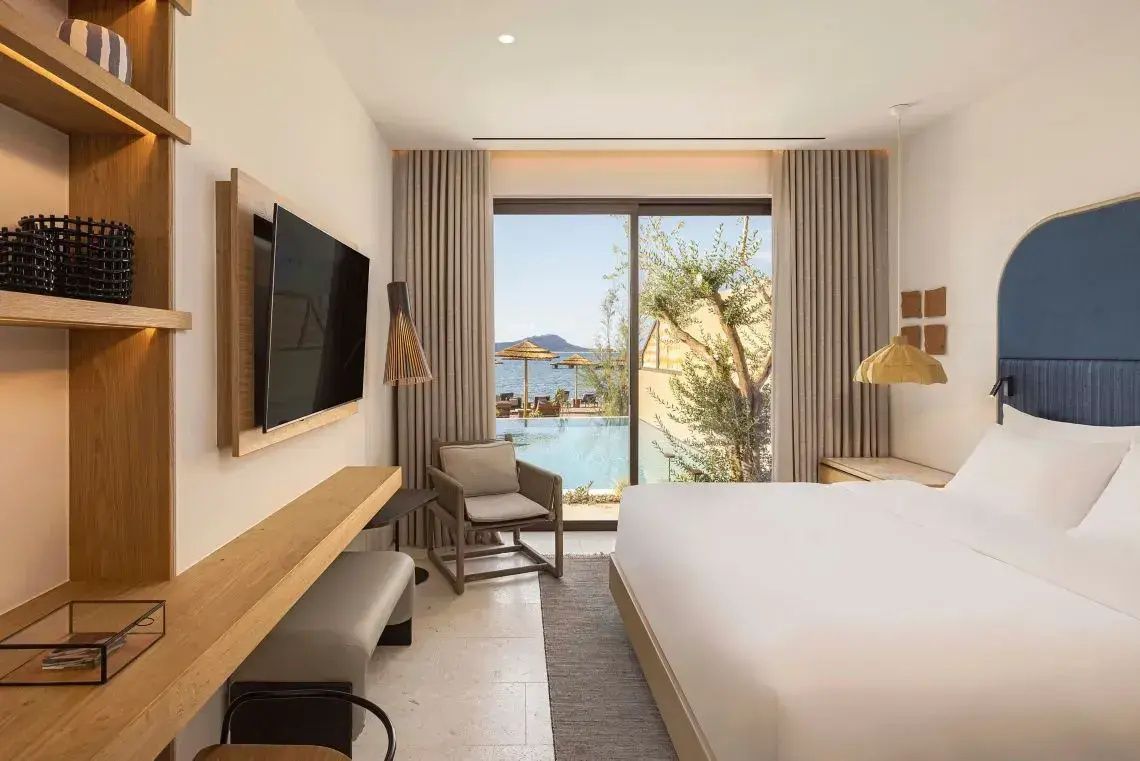 Helles Hotelzimmer mit Doppelbett, Wand-Fernseher und Blick auf die Terrasse mit Pool.