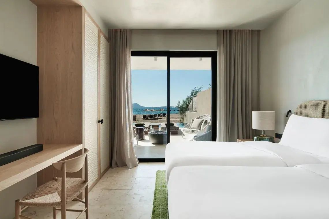 Hotelzimmer mit hellem Bett, Holzschrank und Blick auf die Terrasse am Strand.
