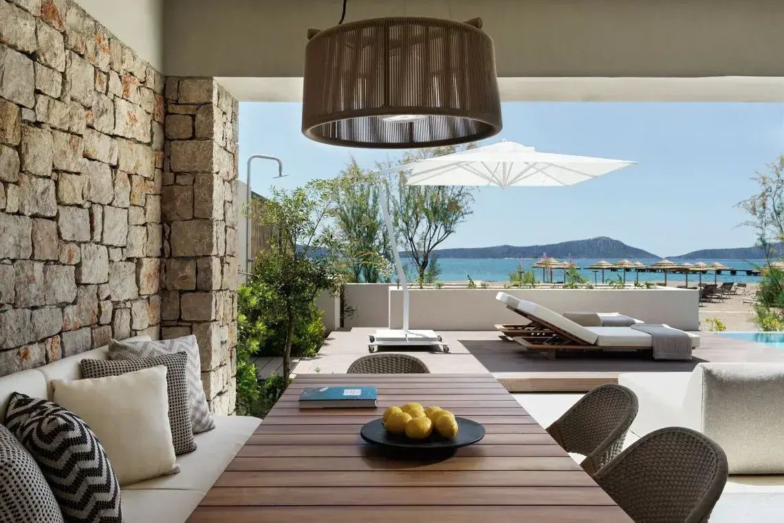 Überdachte Terrasse mit Holztisch, Stühlen und Ausblick auf das Meer und Sonnenschirme.