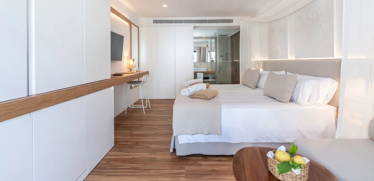 Helles Hotelzimmer mit Doppelbett, Parkettboden, Schreibtisch und angrenzendem Glasbadezimmer.