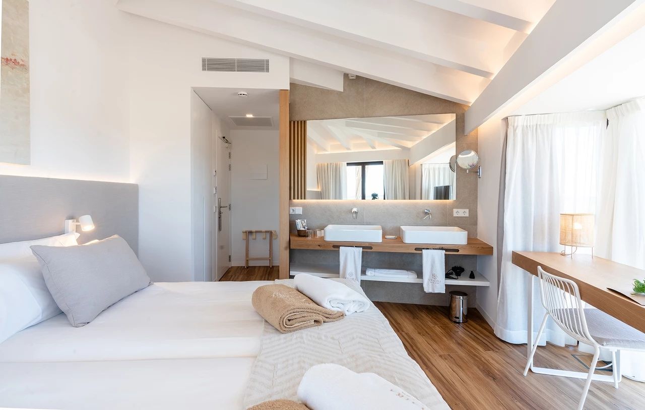 Offenes Hotelzimmer unter einer weißen Holzdecke mit Doppelbett und integriertem Waschbereich.