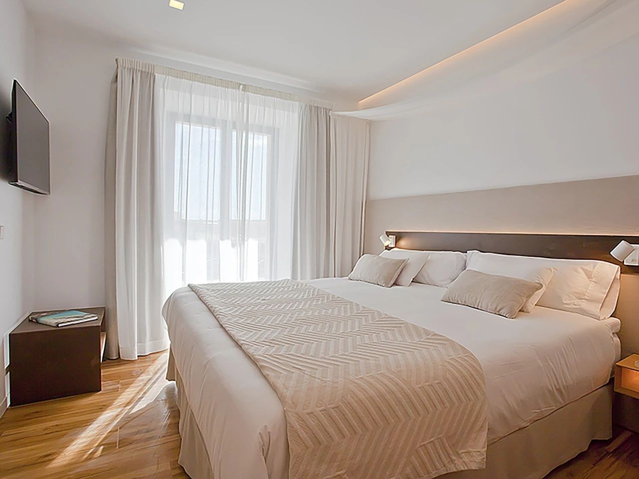 Helles Schlafzimmer mit großem Doppelbett, weißer Bettwäsche und bodentiefen Fenstern hinter Vorhängen.