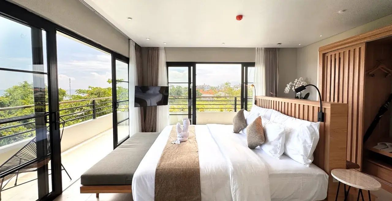 Doppelbett im hellen Hotelzimmer mit großen Fensterfronten und Balkon.