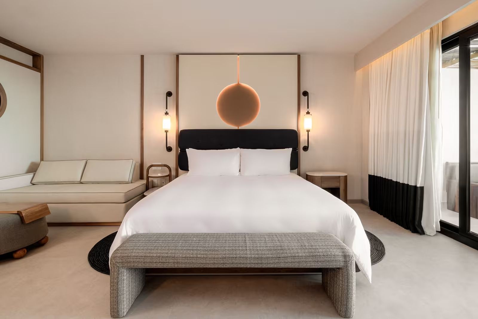 Hell eingerichtetes Hotelzimmer mit großem Doppelbett, gepolsterter Sitzbank und Nachttischlampen.