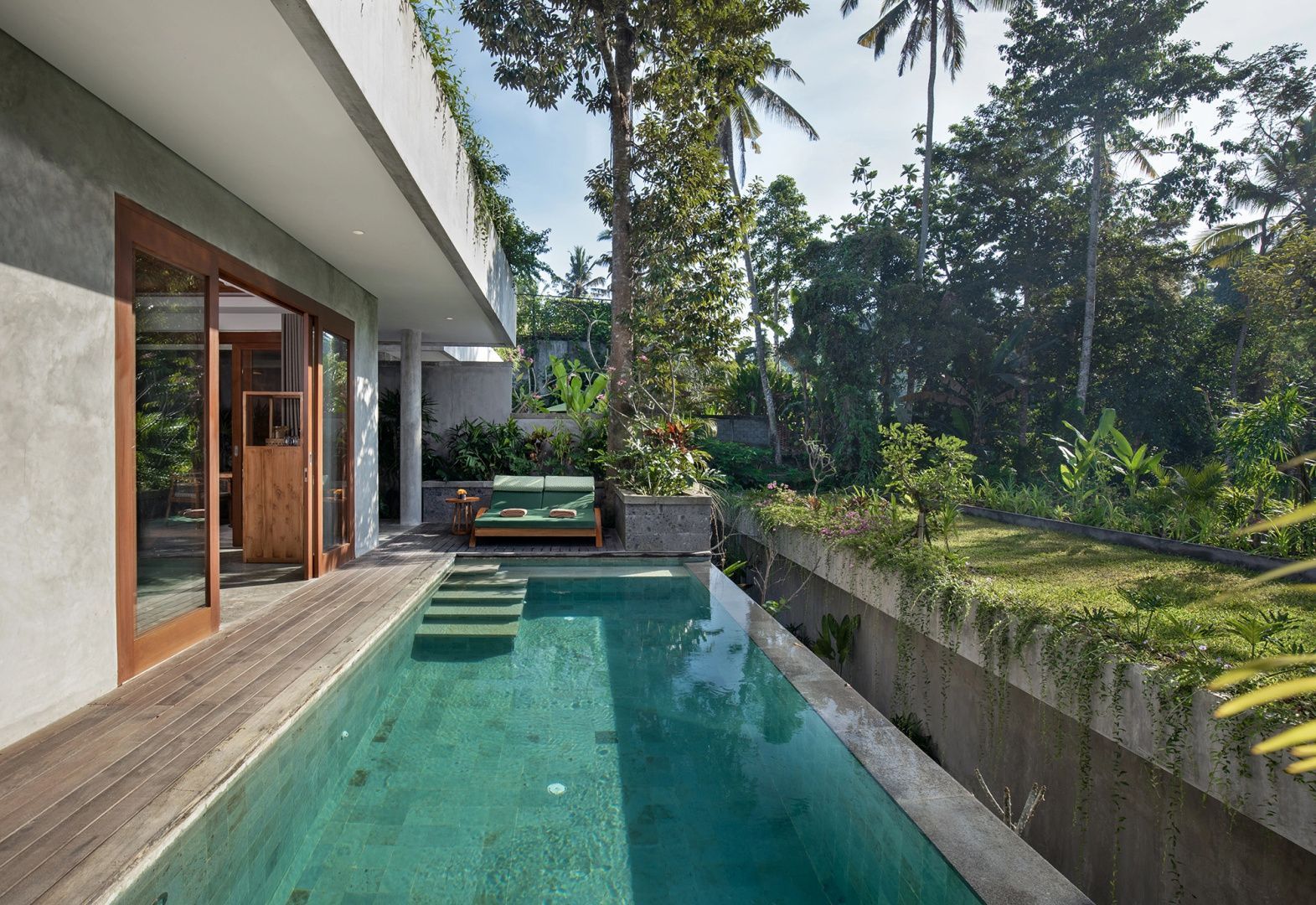 Länglicher Pool auf einer Holzterrasse mit Liegestühlen und von Bäumen umgeben.