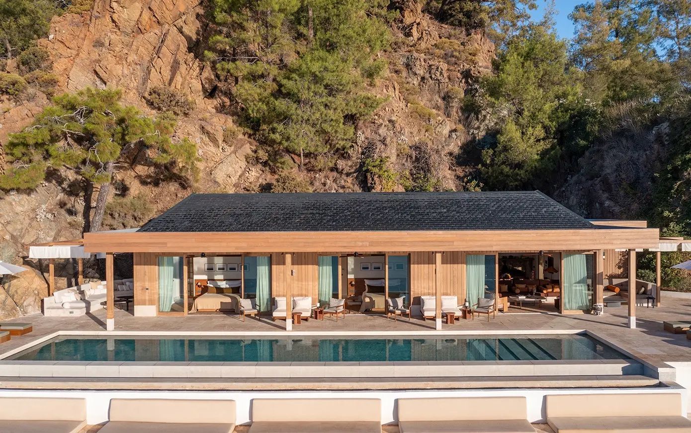Modernes Holzgebäude mit Swimmingpool vor einer steilen Felswand mit Nadelbäumen.