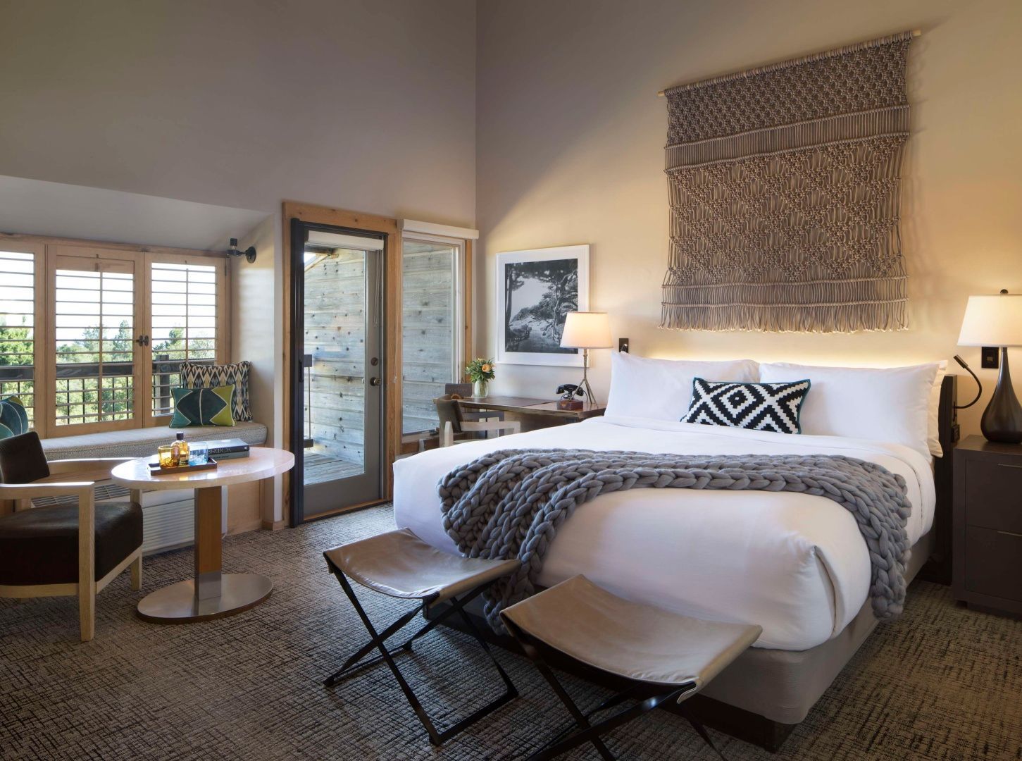 Helles Hotelzimmer mit Doppelbett, Strickdecke, Wandteppich und Zugang zum Balkon.