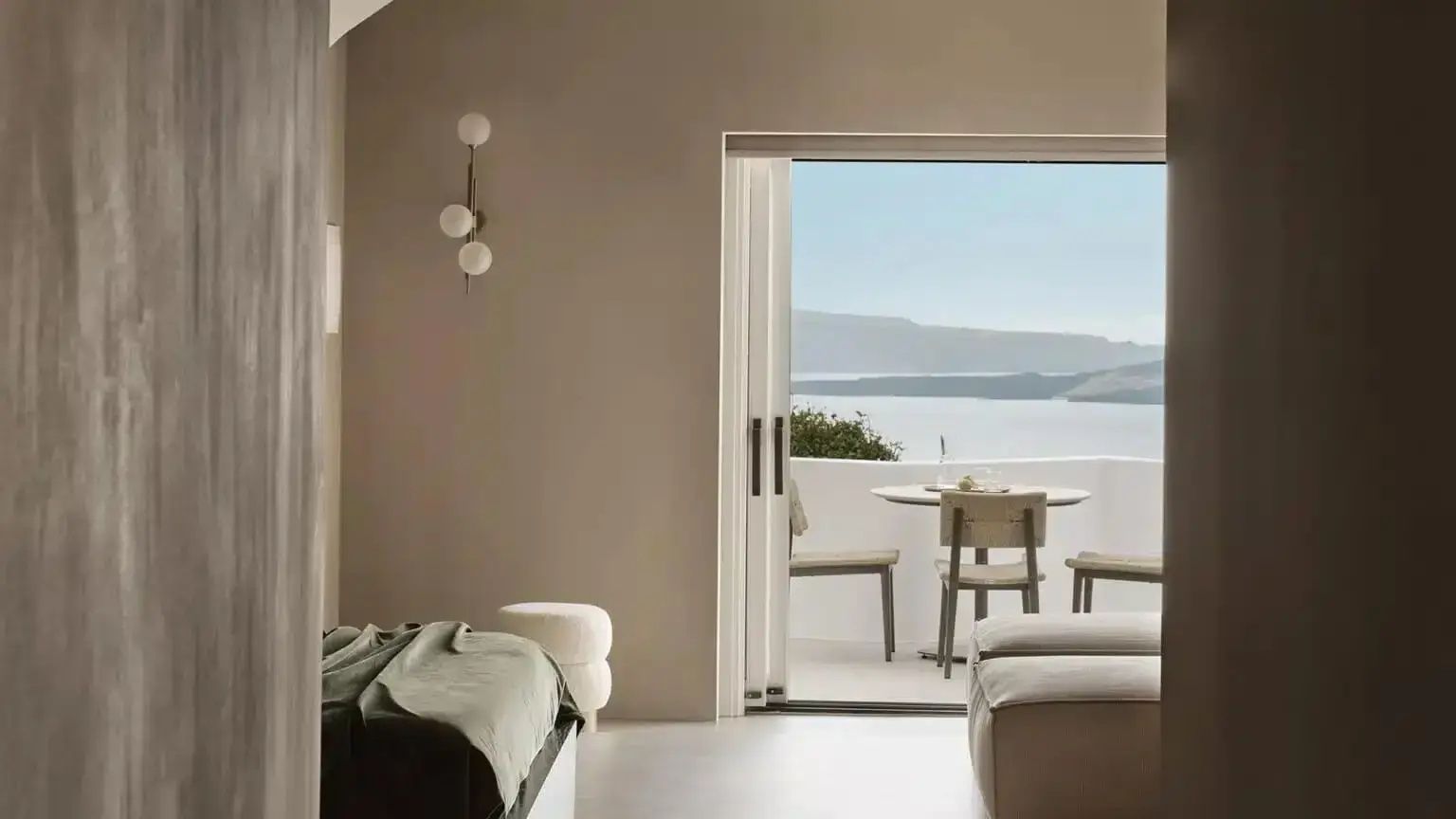 Blick durch die geöffnete Terrassentür auf eine Terrasse mit Sitzgelegenheit und Meerblick.