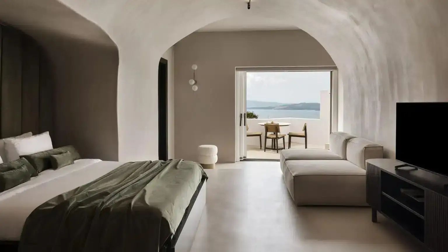 Zimmer in Höhlenarchitektur mit Doppelbett, hellem Sofa und Blick auf den Balkon und das Meer.