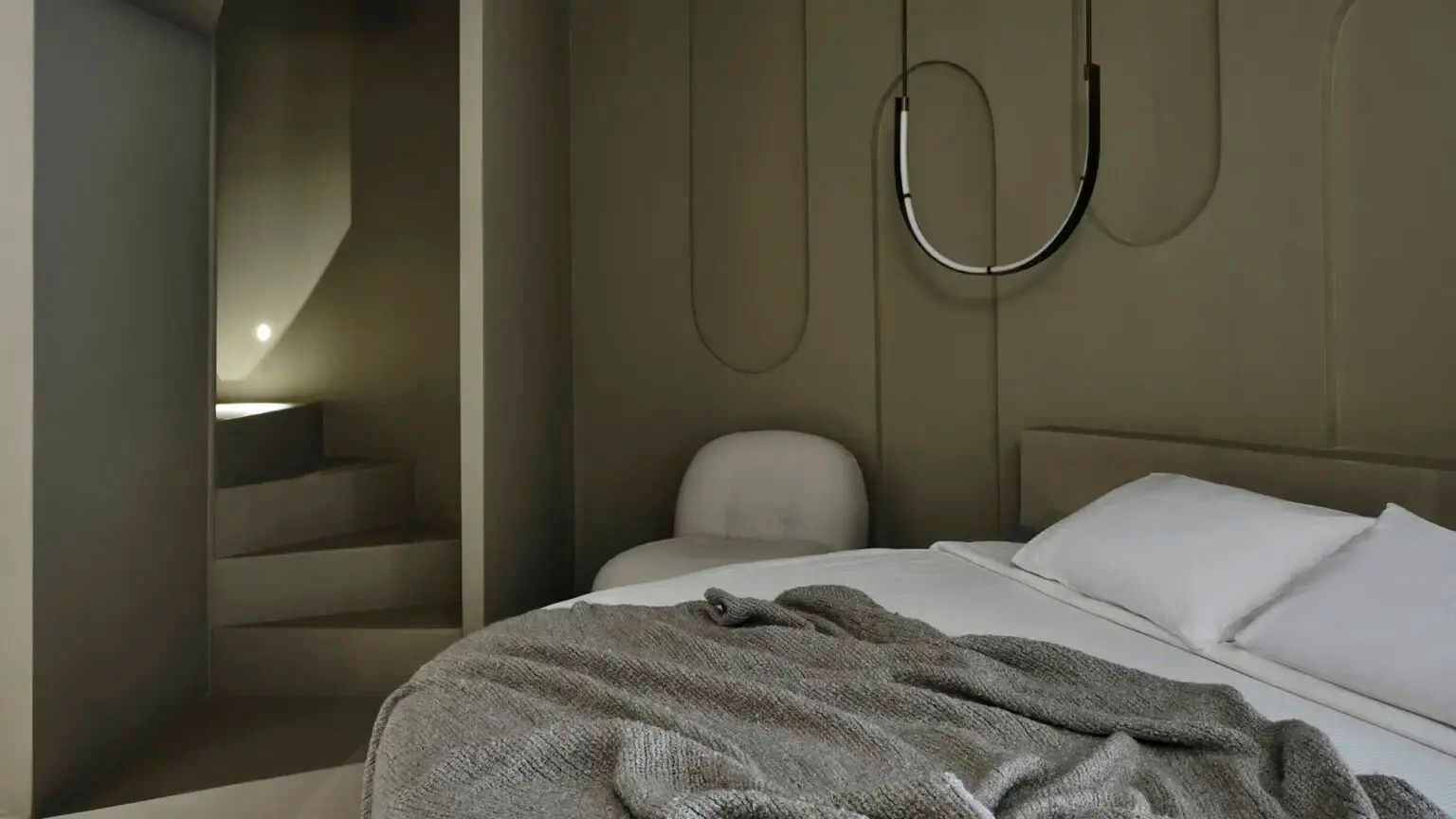 Schlafzimmer mit Doppelbett, hellem Sessel und einer modern geschwungenen Hängelampe an der Wand.