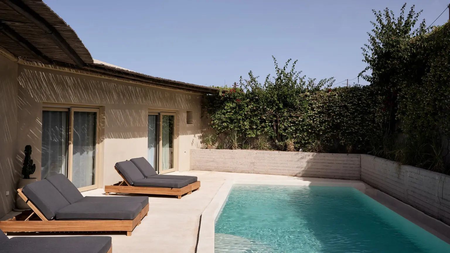 Sonnige Terrasse mit Swimmingpool, zwei Sonnenliegen aus Holz und einer bepflanzten Steinmauer.