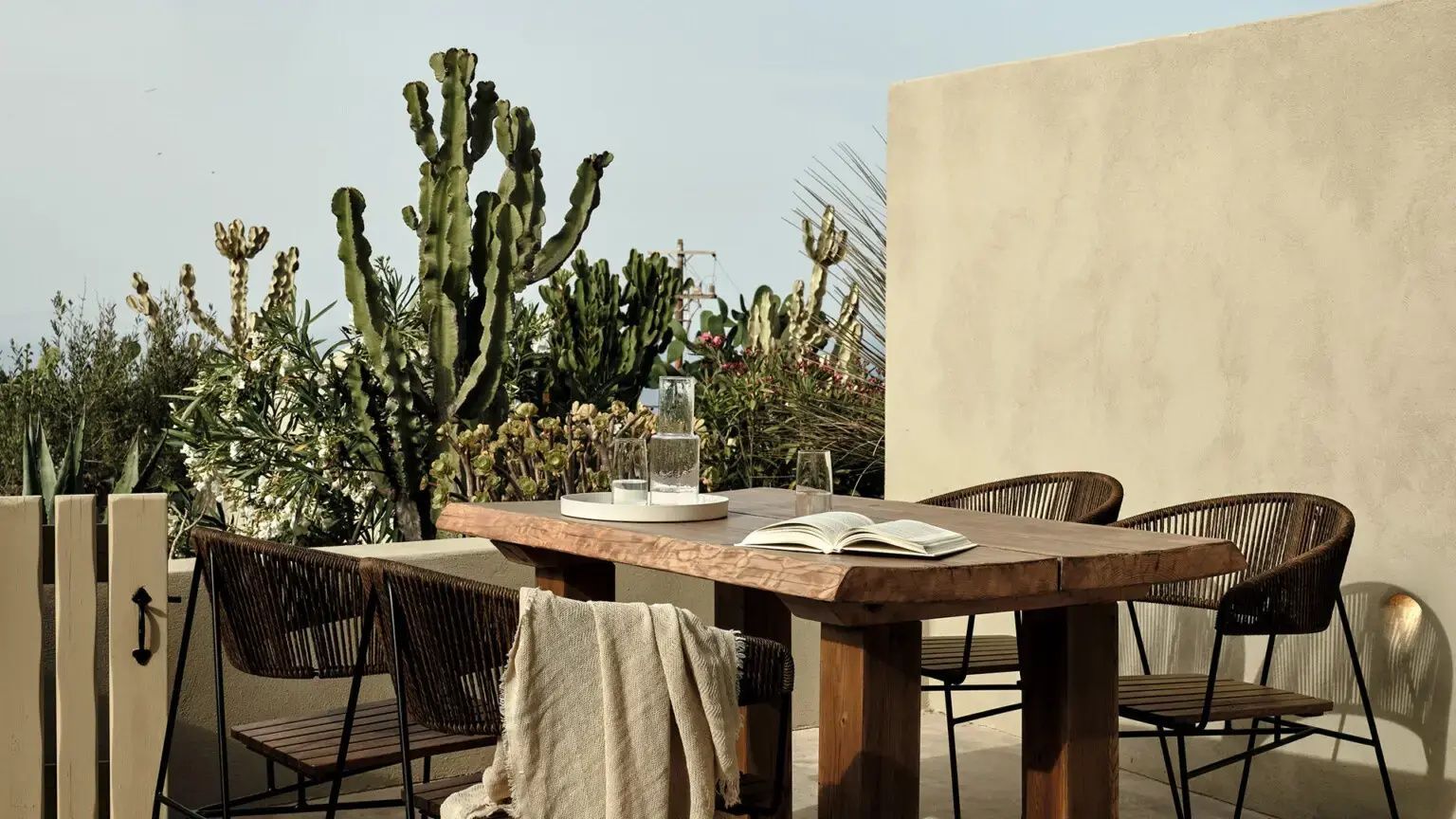 Holztisch mit Stühlen auf einer Sonnenterrasse, umgeben von Kakteen und mediterranen Pflanzen.