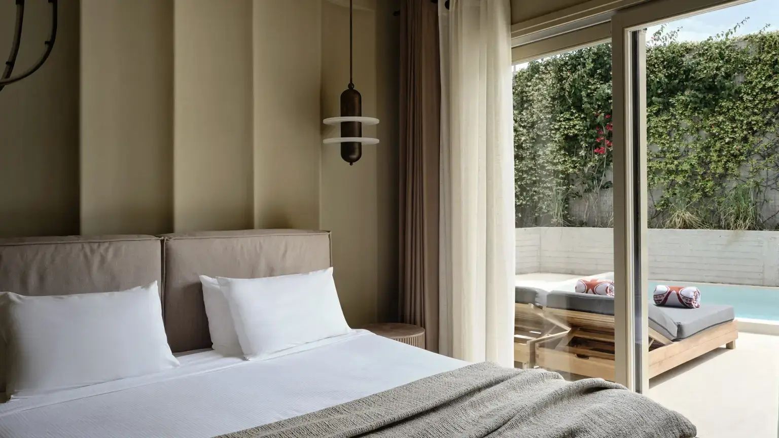 Schlafzimmer mit weiß gedecktem Bett, Vorhängen und Durchgang zur Terrasse mit Liegen.