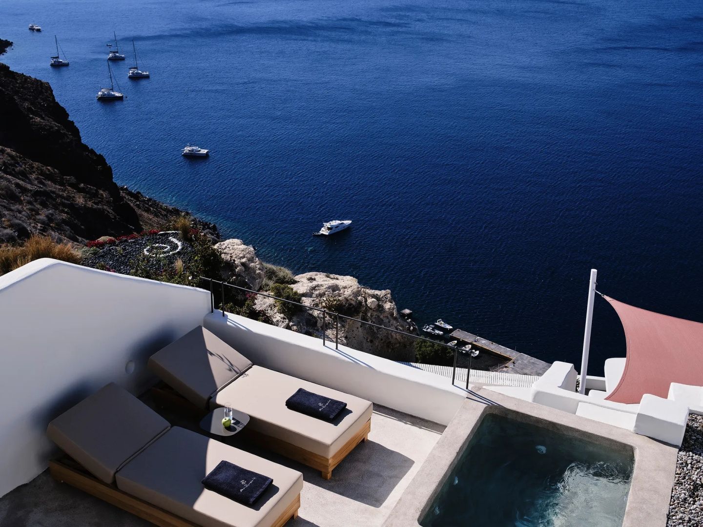 Blick von oben auf eine Terrasse mit Liegen, Whirlpool, steiler Felswand und das blaue Meer.
