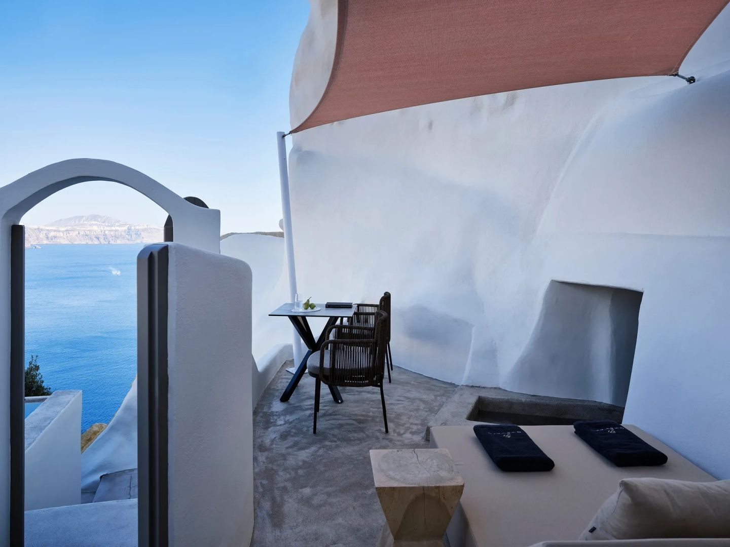 Terrasse an weißer Felswand mit Tisch, Stühlen und Ausblick auf das Meer und die Küste von Santorini.