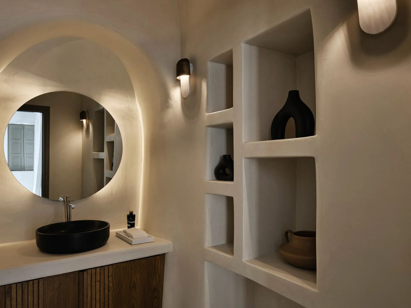 Badezimmer mit beleuchtetem rundem Spiegel, schwarzem Waschbecken und Wandnischen.