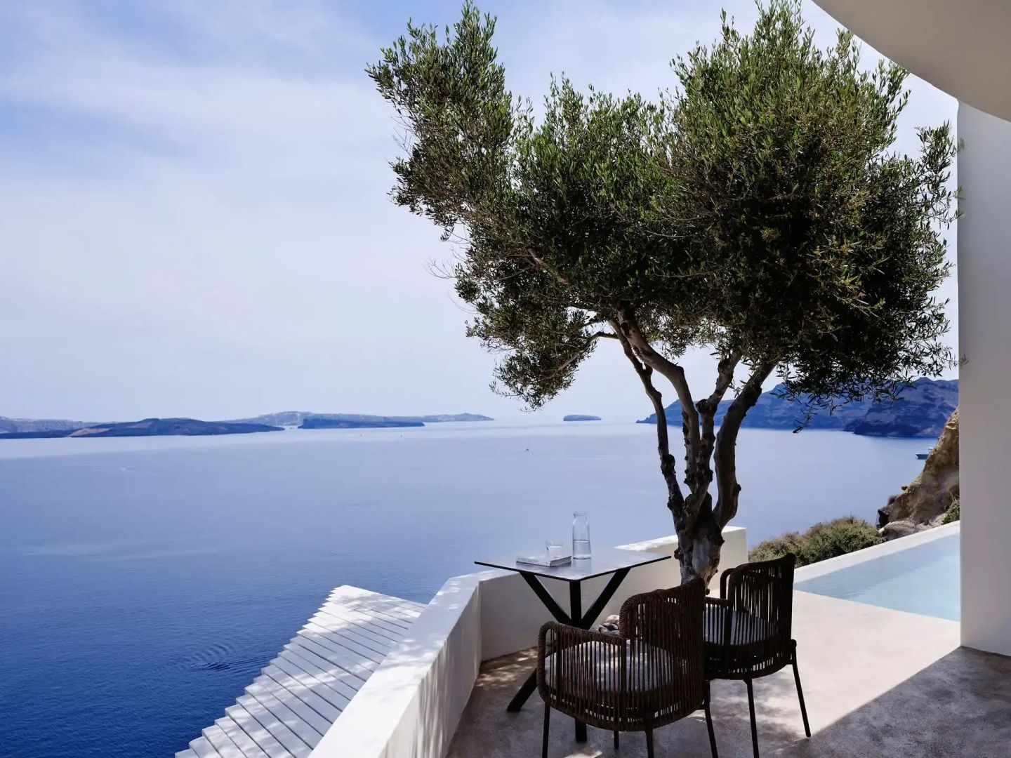 Terrasse mit Sitzgelegenheiten unter einem Olivenbaum und Blick auf das blaue Meer.
