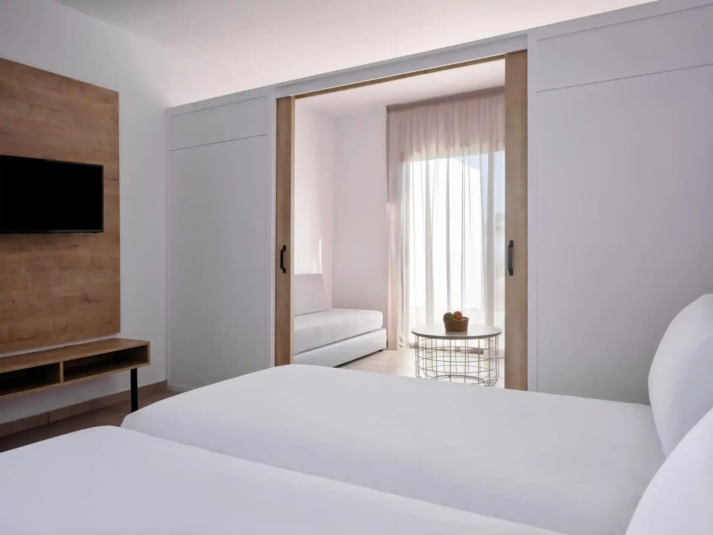 Helles Hotelzimmer mit Doppelbett und Blick durch eine Schiebetür in den Wohnbereich.