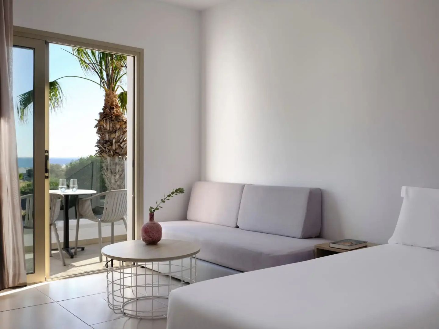 Sitzbereich im Hotelzimmer mit weißem Sofa, Tisch und Blick auf den Balkon.