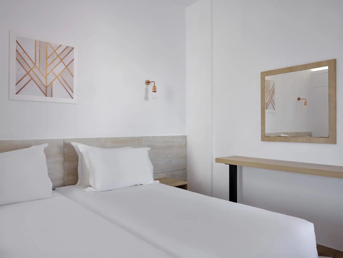 Schlafzimmer mit weißem Bett, Holzablage und einem Wandspiegel an weißer Wand.