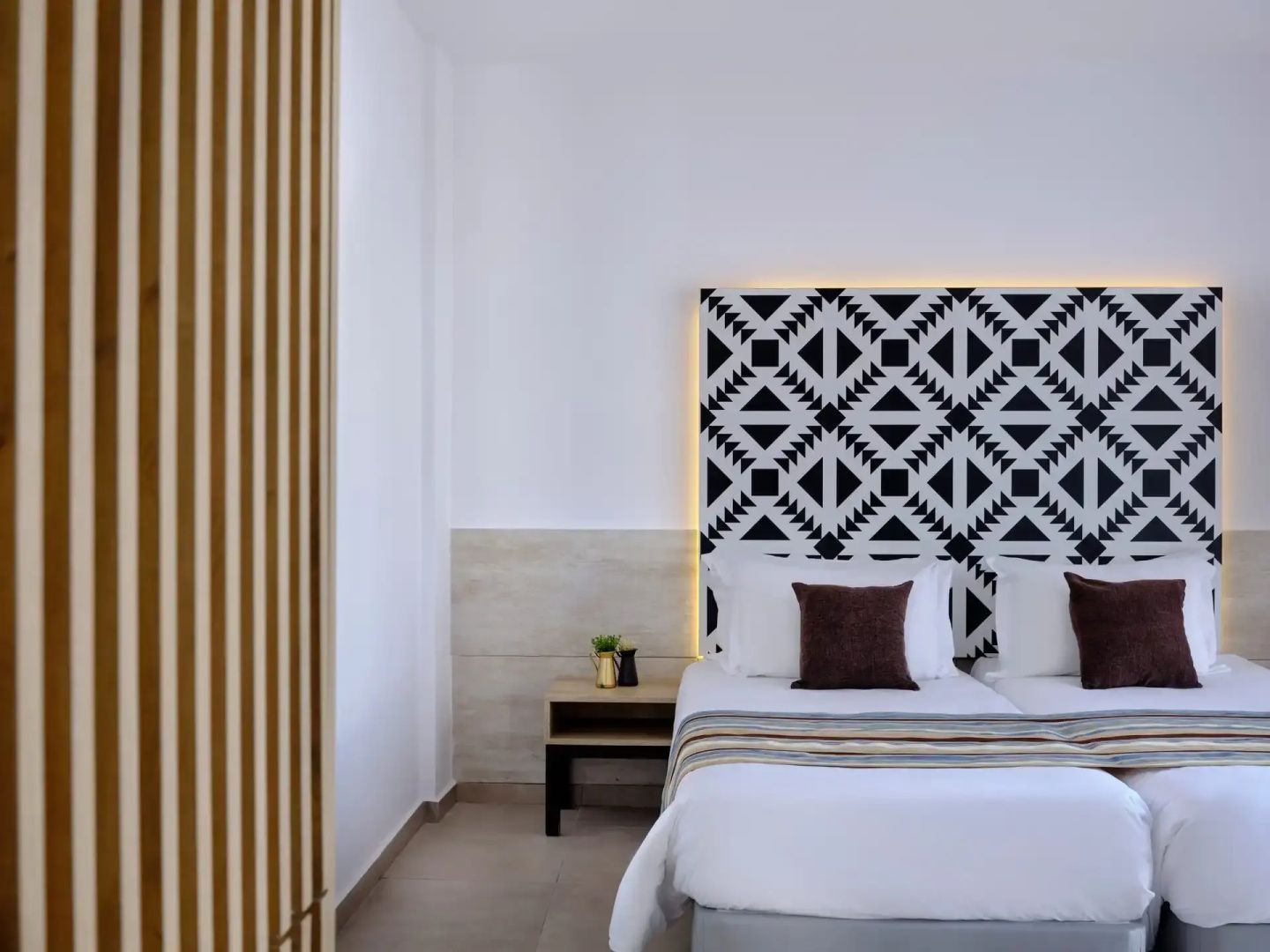 Moderne Betten im Hotelzimmer mit gemustertem Kopfteil, weißer Bettwäsche und hölzernem Nachttisch.