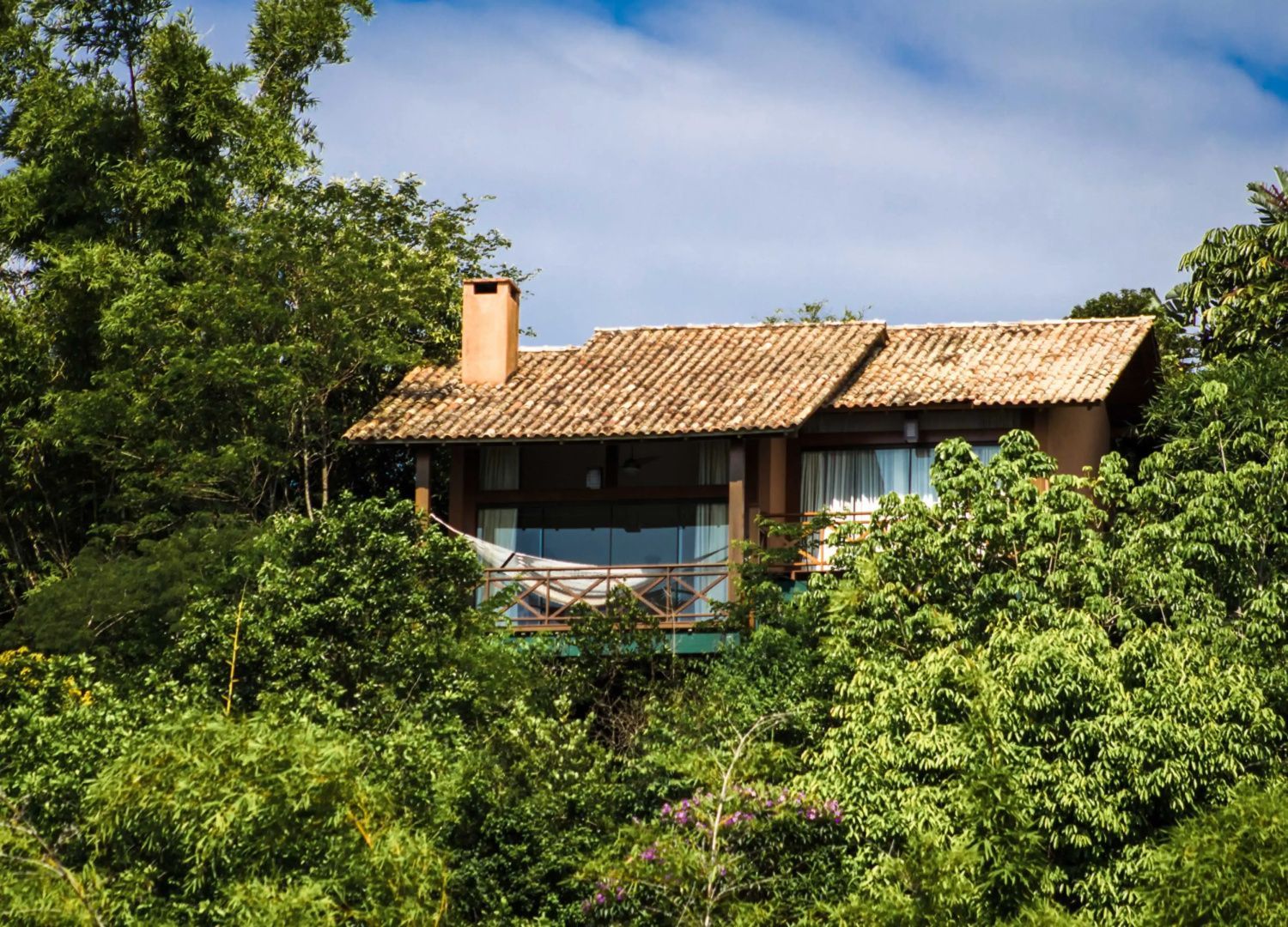 Bungalow aus Holz mit Ziegeldach und Hängematte auf dem Balkon im Grünen.