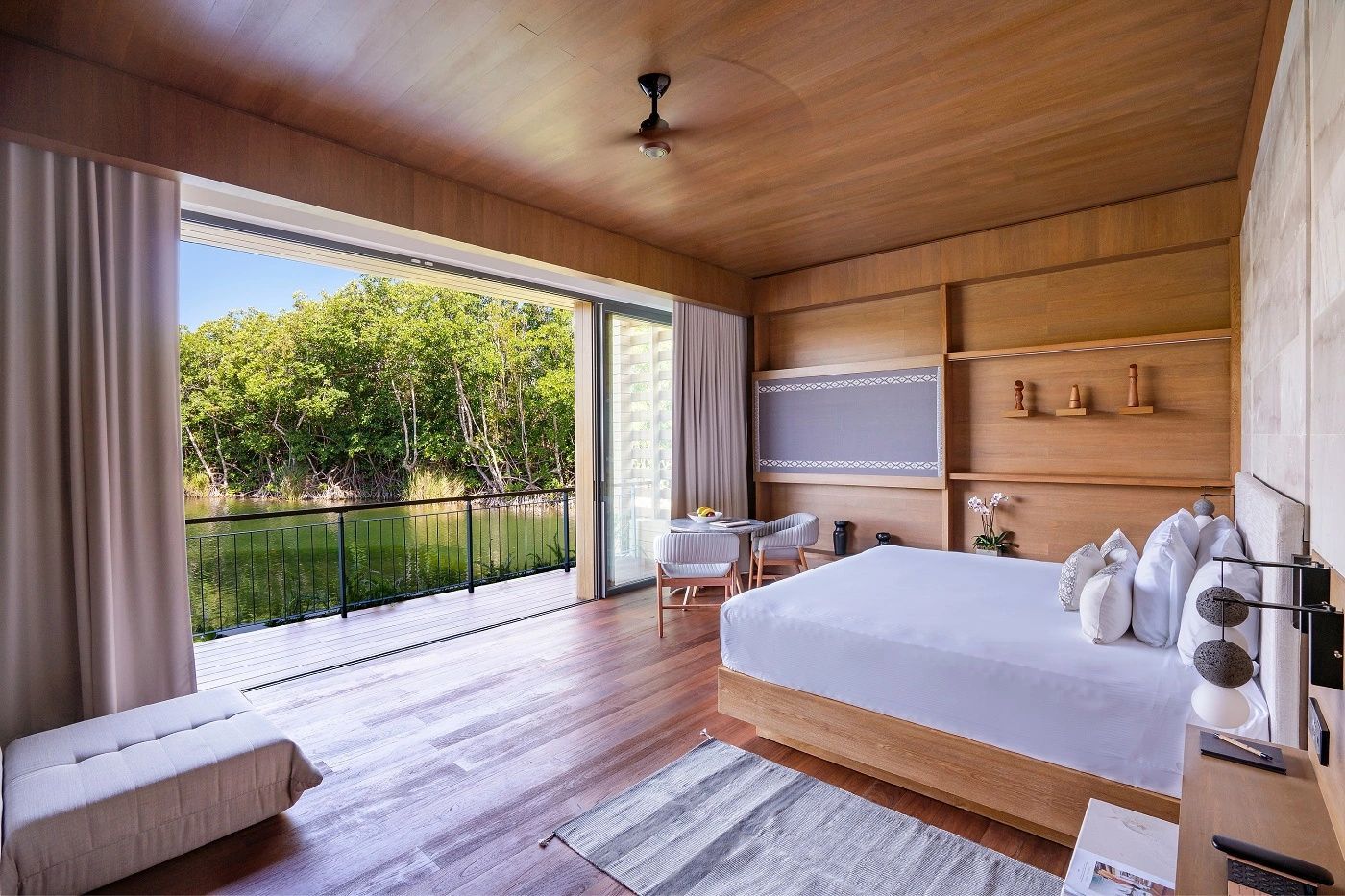 Helles Hotelzimmer mit Doppelbett, Holzwänden und geöffneter Front zu einer Terrasse am Wasser.