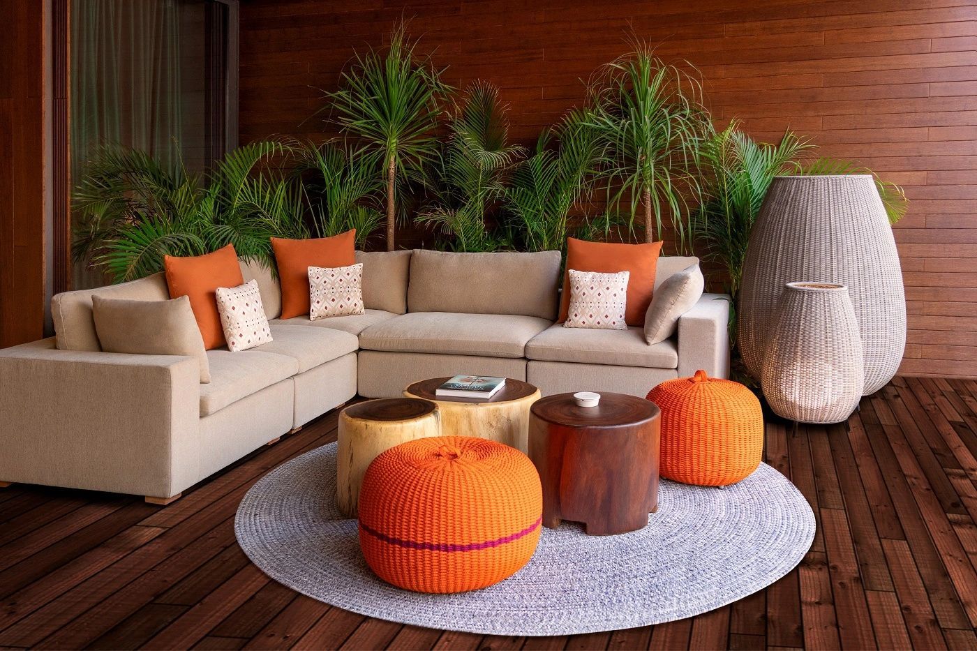 Sitzgelegenheit auf einer Holzterrasse mit beigefarbenem Ecksofa, orangen Kissen und Hockern.