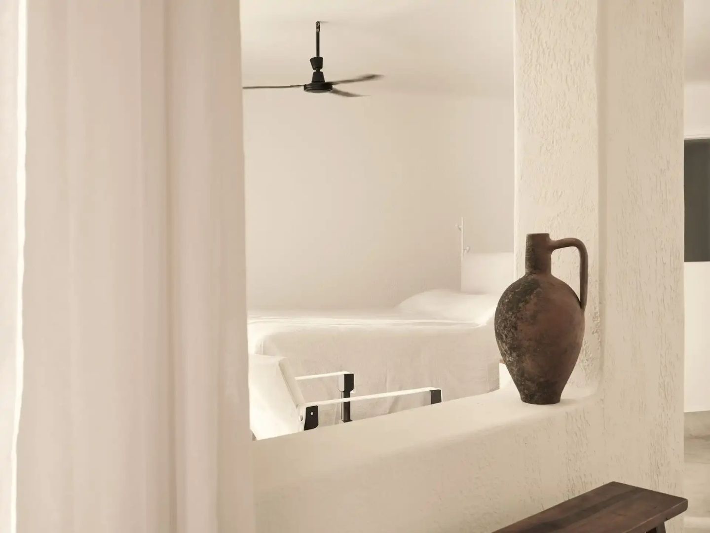 Blick durch einen Wanddurchbruch auf ein weißes Doppelbett mit Deckenventilator und Tonkrug im Vordergrund.