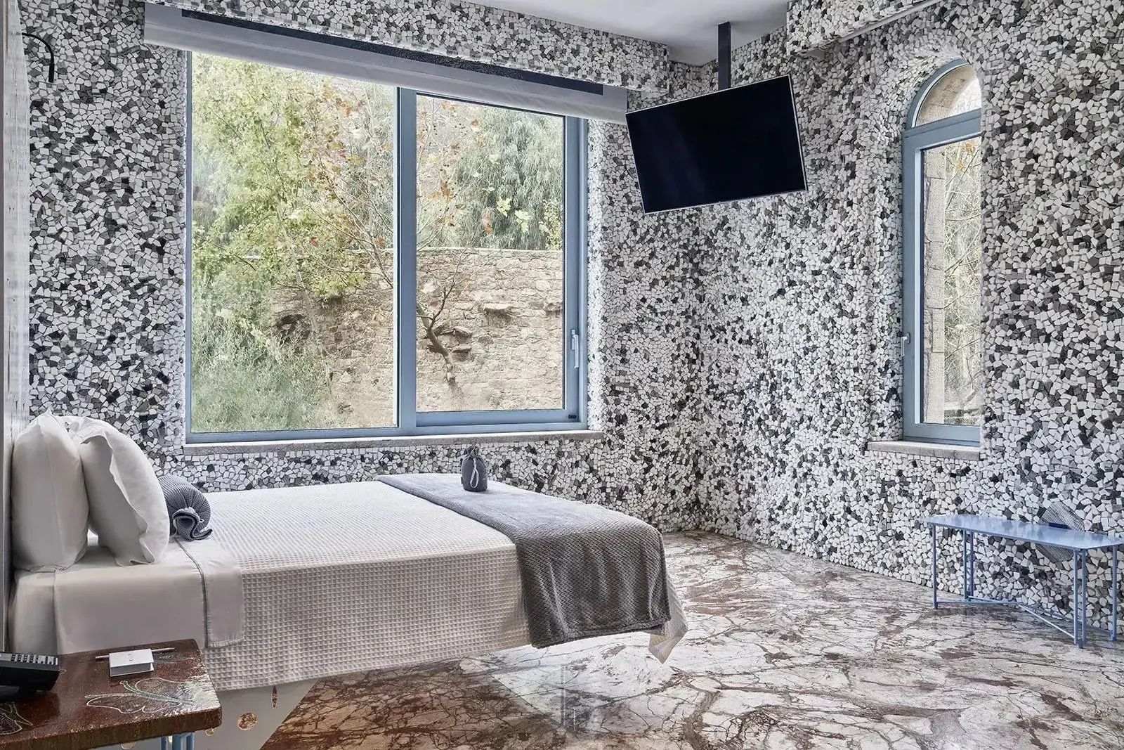 Hotelzimmer mit grau-weißer Mosaikwand, großem Fenster und weiß bezogenem Bett.