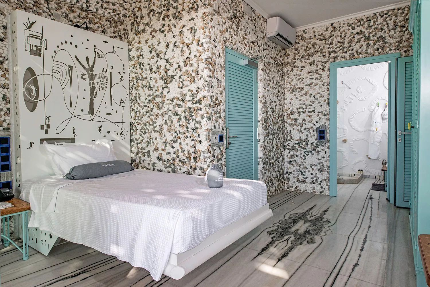 Schlafbereich mit weiß bezogenem Bett, kunstvoll gemusterter Steinwand und blauen Türen.