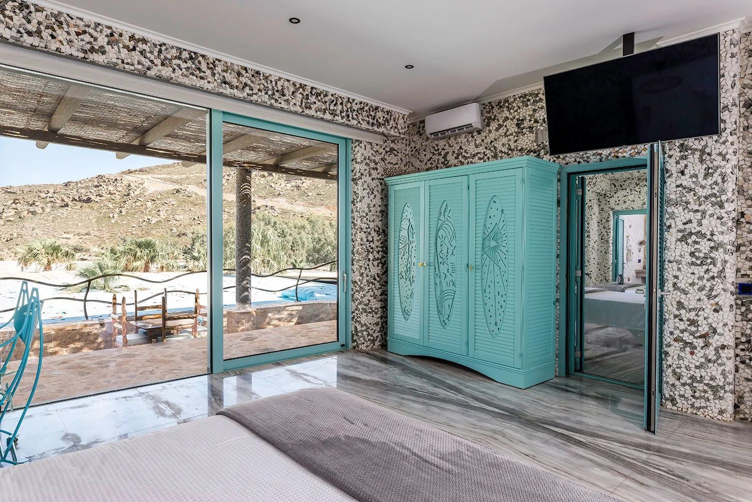 Schlafzimmer mit türkisblauem Schrank, Marmorboden und Fenstern zur Terrasse.