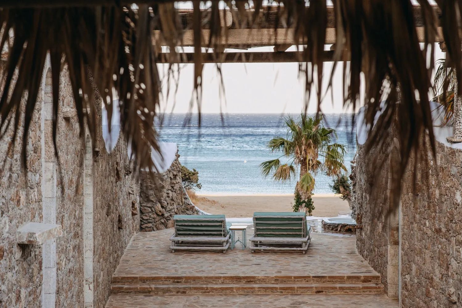 Blick durch eine überdachte Terrasse auf Liegestühle, Strand und das Meer.