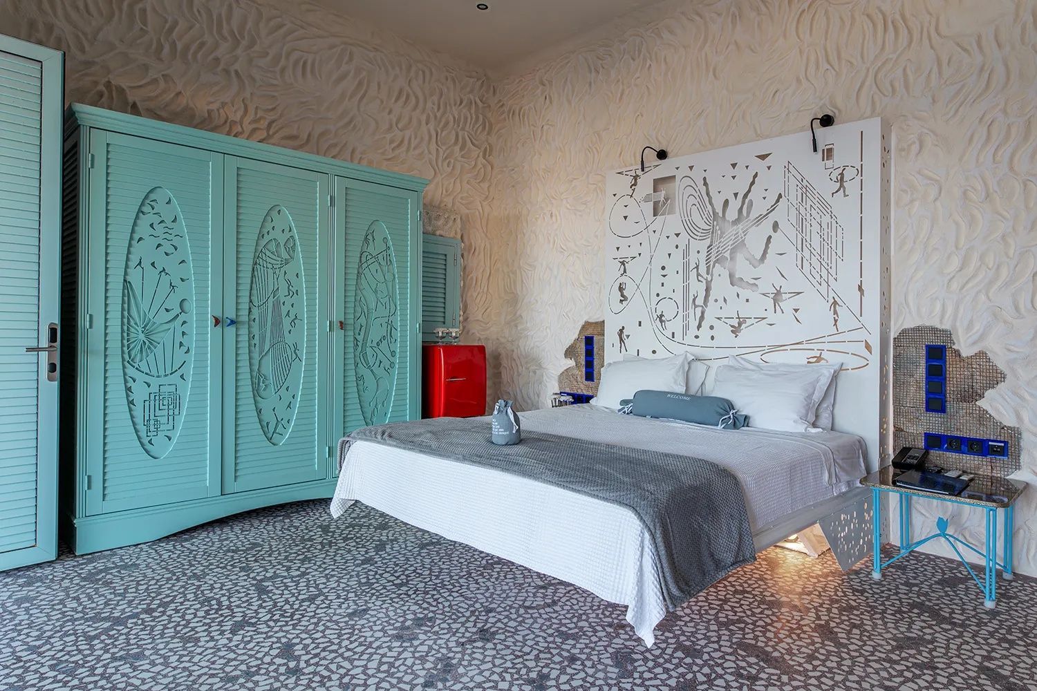 Doppelbett mit kunstvollem Kopfteil, türkisblauem Schrank und rotem Kühlschrank.