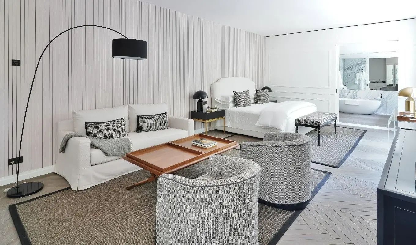 Hell gestaltetes Zimmer mit Bett, weißem Sofa, zwei Sesseln und Holztisch.