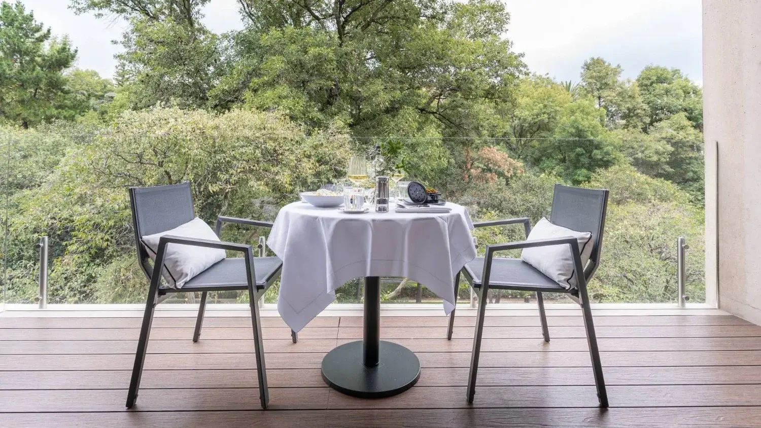 Gedeckter Tisch mit zwei Stühlen auf einem Balkon mit Blick auf dichte grüne Bäume.