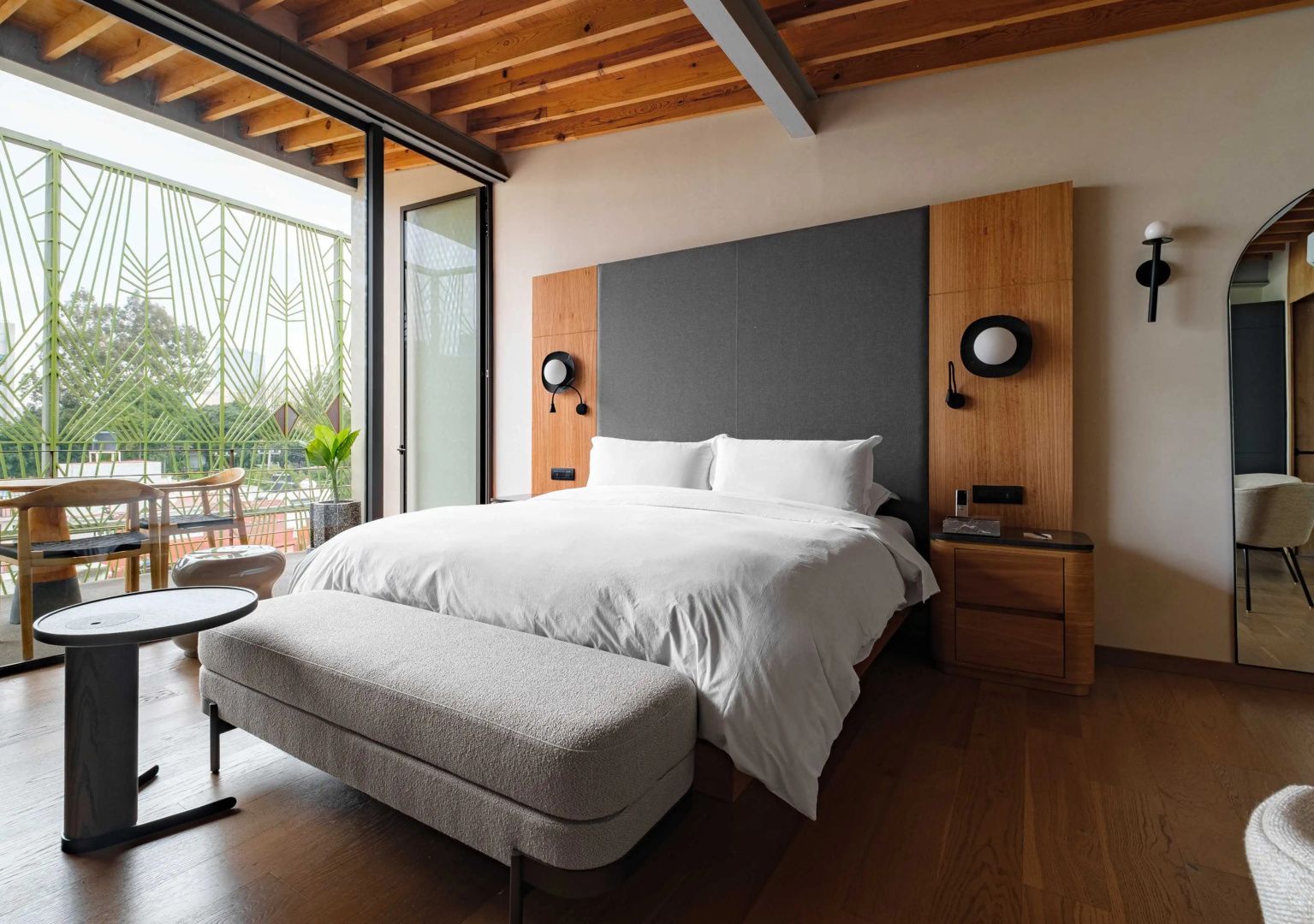 Doppelbett mit Holzpaneel an der Wand und gepolsterter Bank im hellen Hotelzimmer mit Balkonzugang.