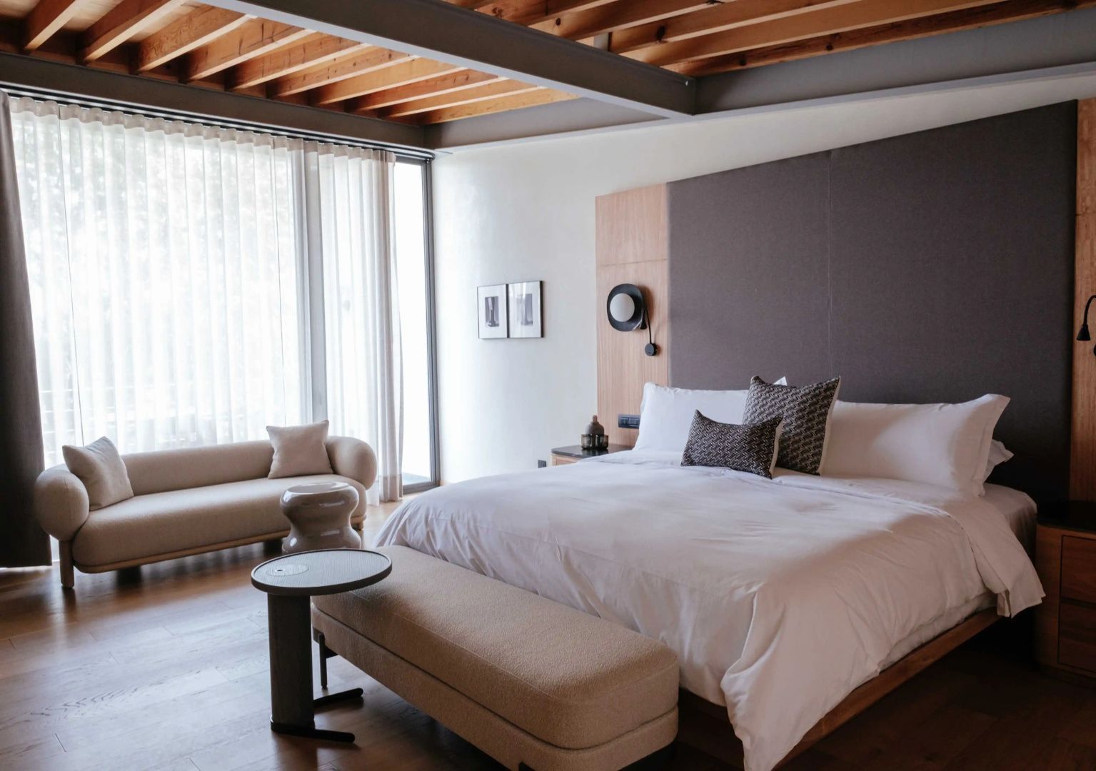 Doppelbett mit weißer Bettwäsche, Holzdecke und Sitzcouch vor großem Fenster.