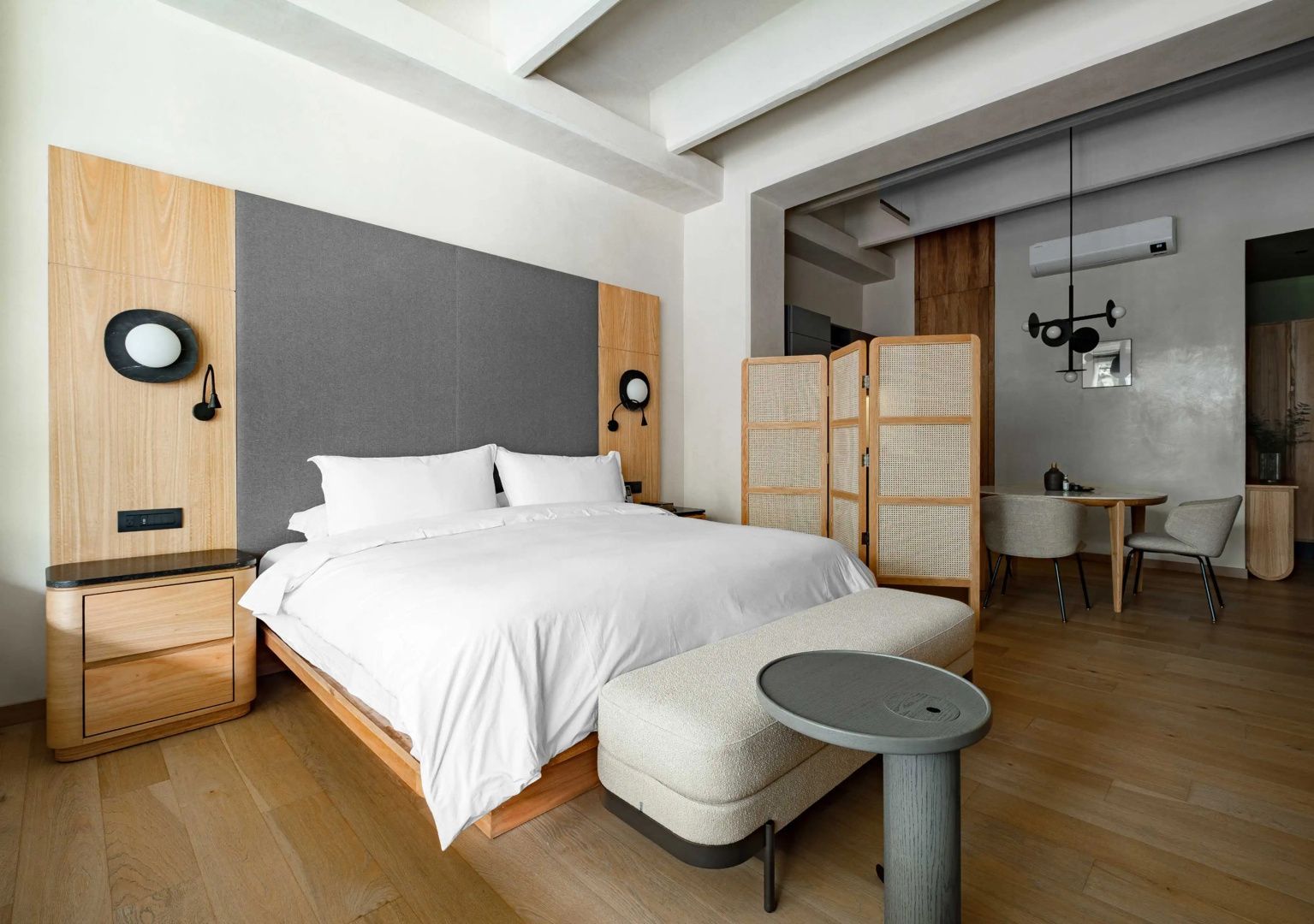 Großes Doppelbett mit grauem Kopfteil, Parkettboden und einem Paravent aus Geflecht.
