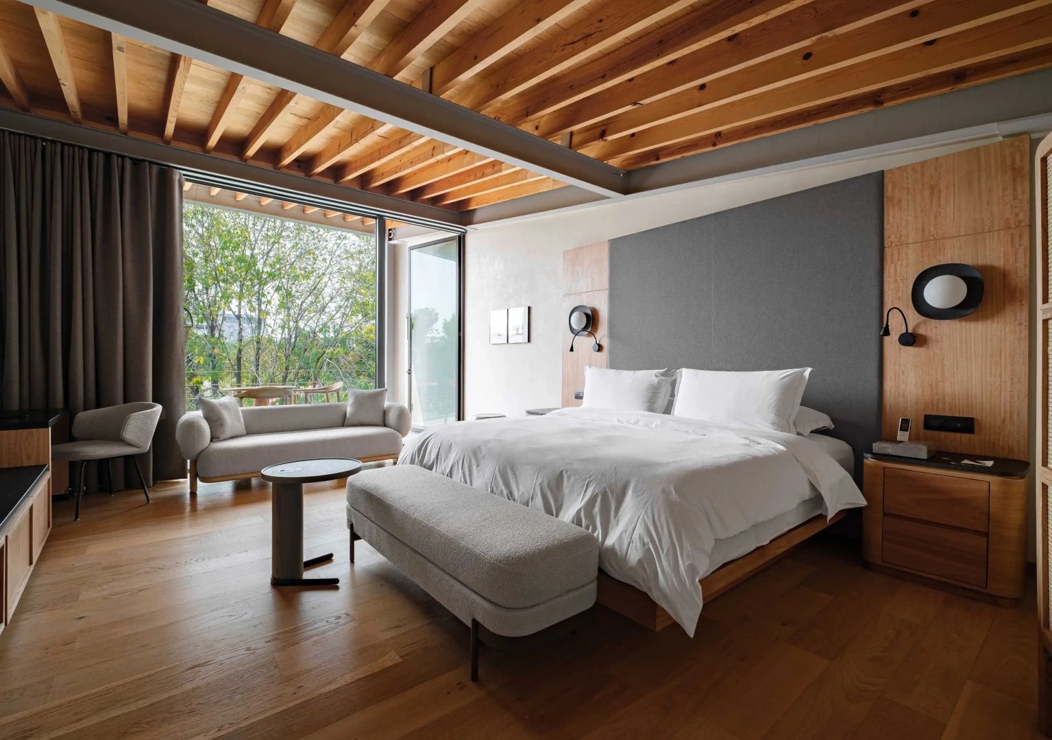 Geräumiges Hotelzimmer mit Parkettboden, Holzbalkendecke, Doppelbett und Sitzcouch vor großem Fenster.