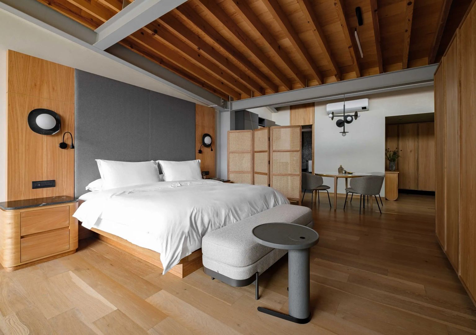 Hotelzimmer mit Holzbalkendecke, Bett, flexiblem Raumteiler und Esstisch mit Stühlen im Hintergrund.