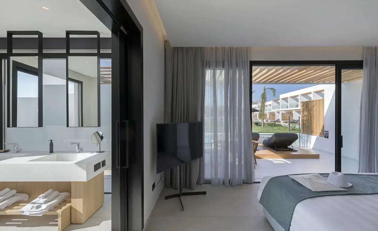 Moderner Hotelraum mit Fernseher, Blick ins Bad und Zugang zur Terrasse mit Sonnenliegen.