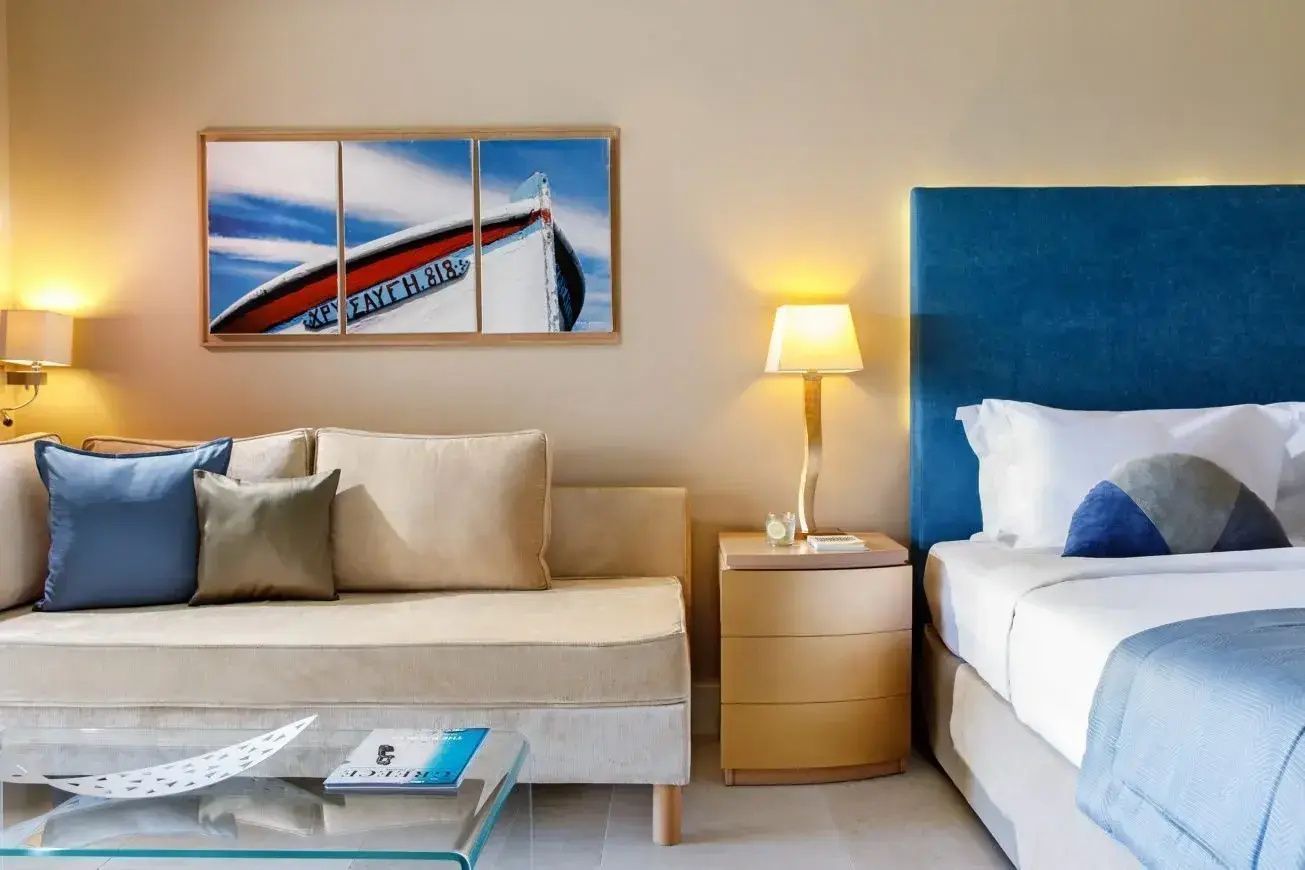 Helles Hotelzimmer mit beigem Sofa, Nachttisch mit Lampe, blauem Kopfteil und Wandbild eines Bootes.