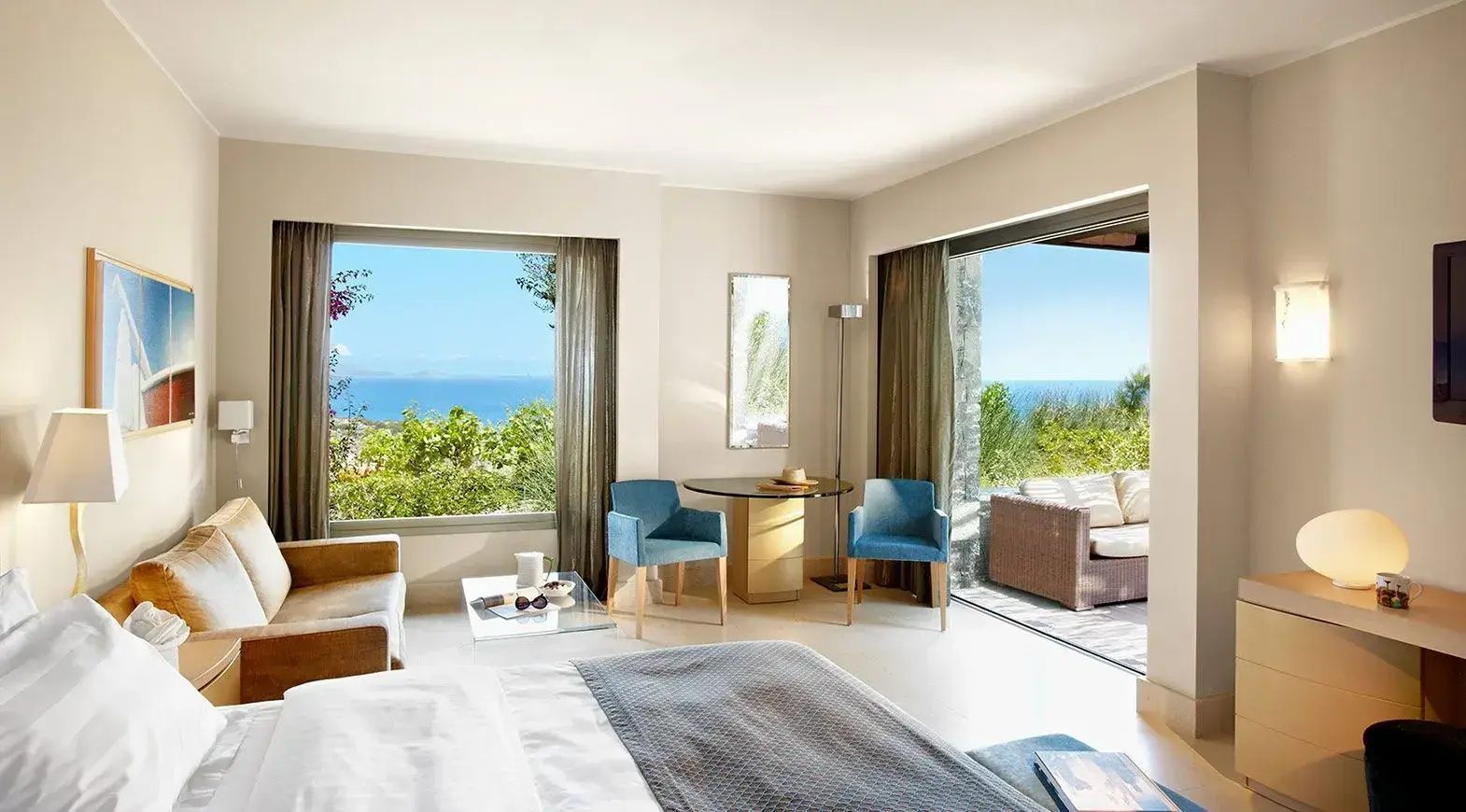 Helles Hotelzimmer mit Betten, zwei blauen Sesseln und Ausblick auf das Meer.