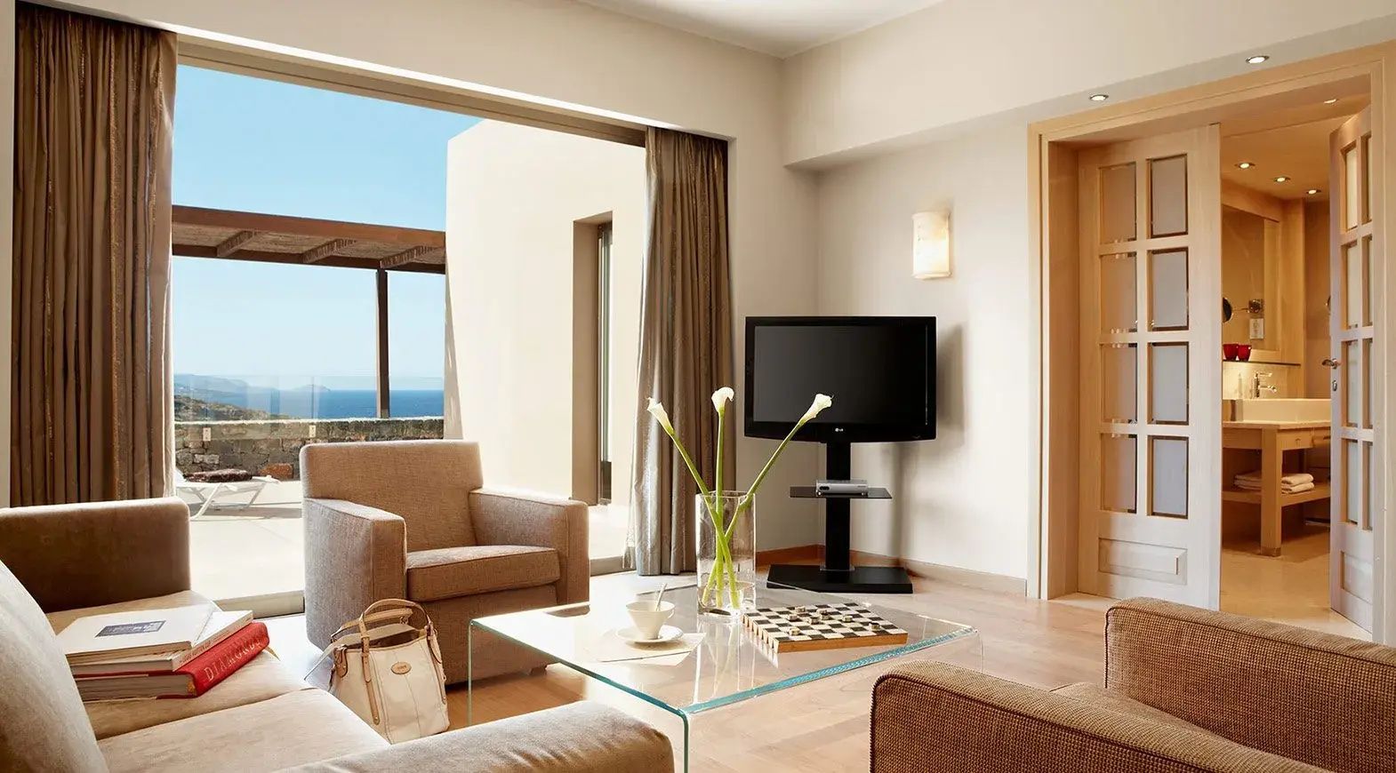 Helles Wohnzimmer mit Sesseln, Glastisch, Fernseher und Balkonzugang.