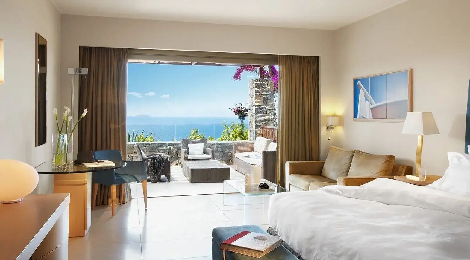 Helles Hotelzimmer mit Doppelbett, Sofa und großer Glasfront zur Terrasse mit Meerblick.