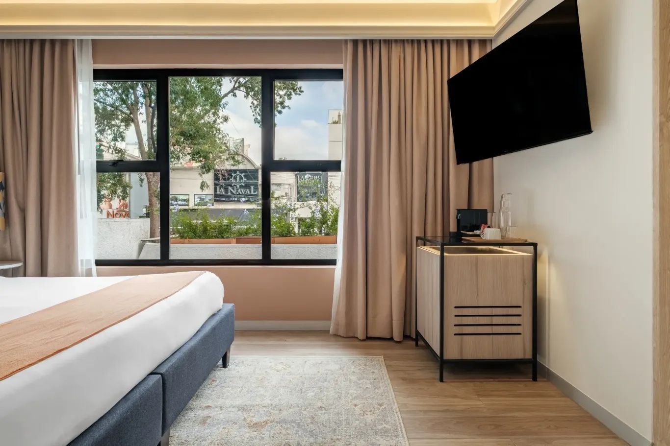 Hotelzimmer mit Fensterfront, Flachbild TV an der Wand und kleiner Anrichte.