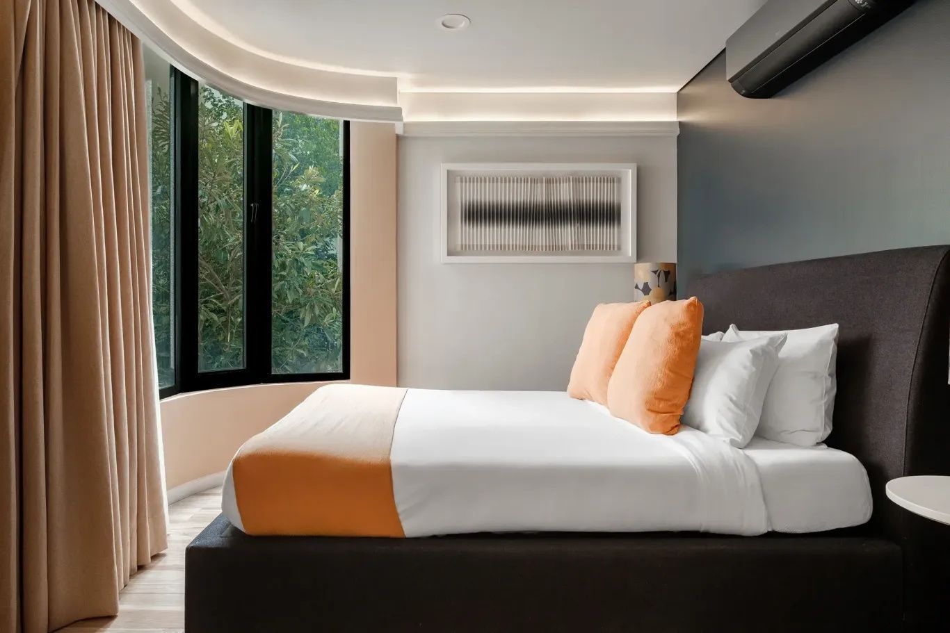 Doppelbett mit orangefarbenen Kissen vor einer großen Fensterfront mit Blick ins Grüne.
