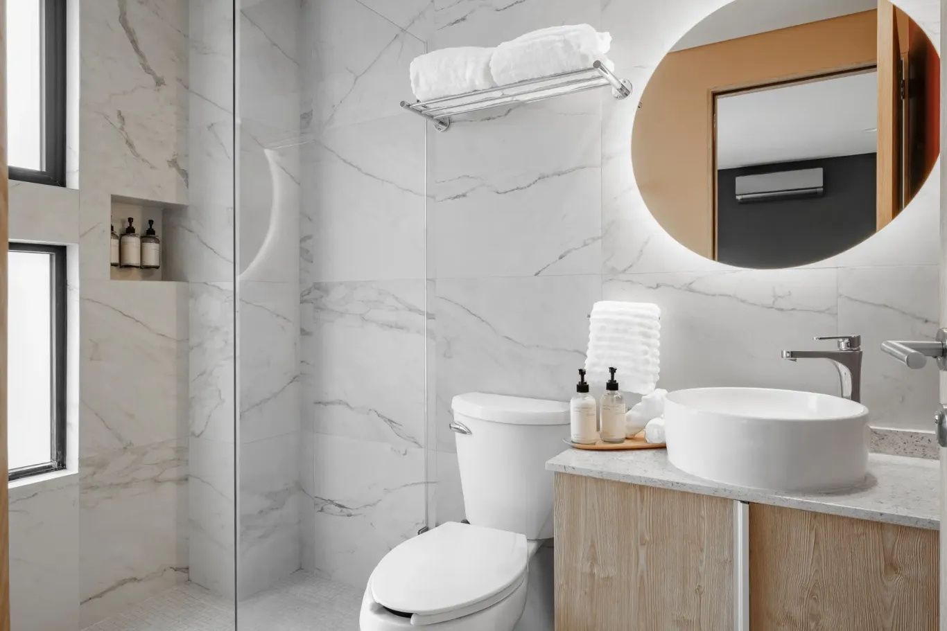 Badezimmer mit weißer Marmoroptik, Glasdusche, Waschtisch und rundem Spiegel.