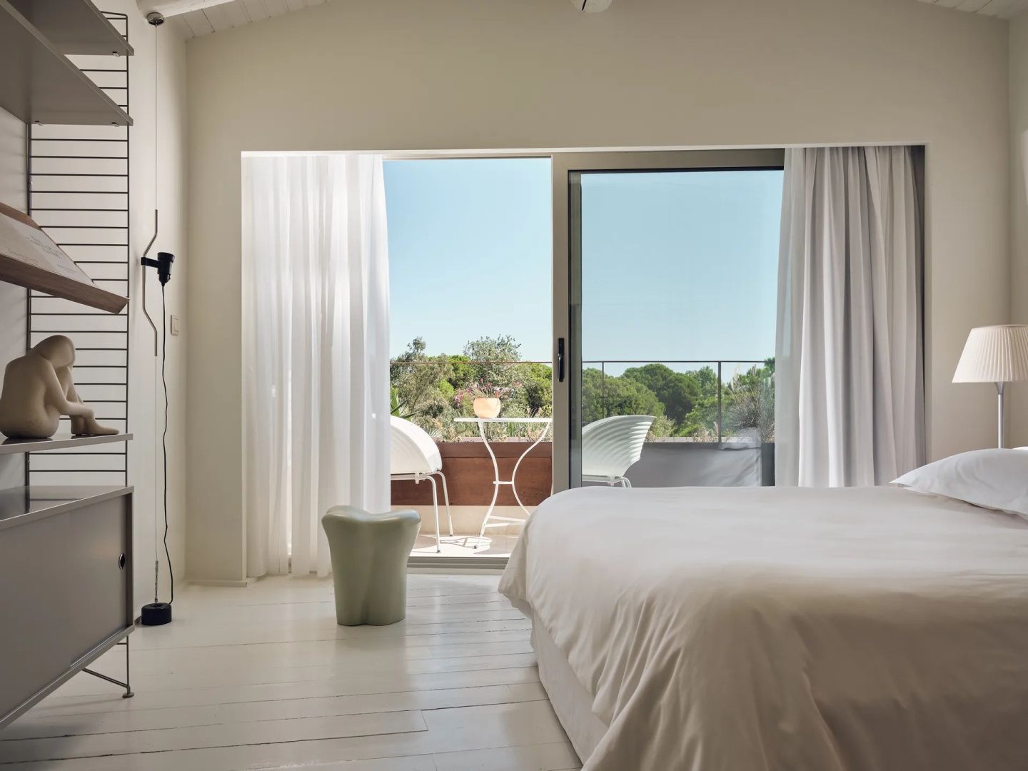 Helles Schlafzimmer mit weißem Bett und geöffneter Schiebetür zum Balkon mit Blick ins Grüne.